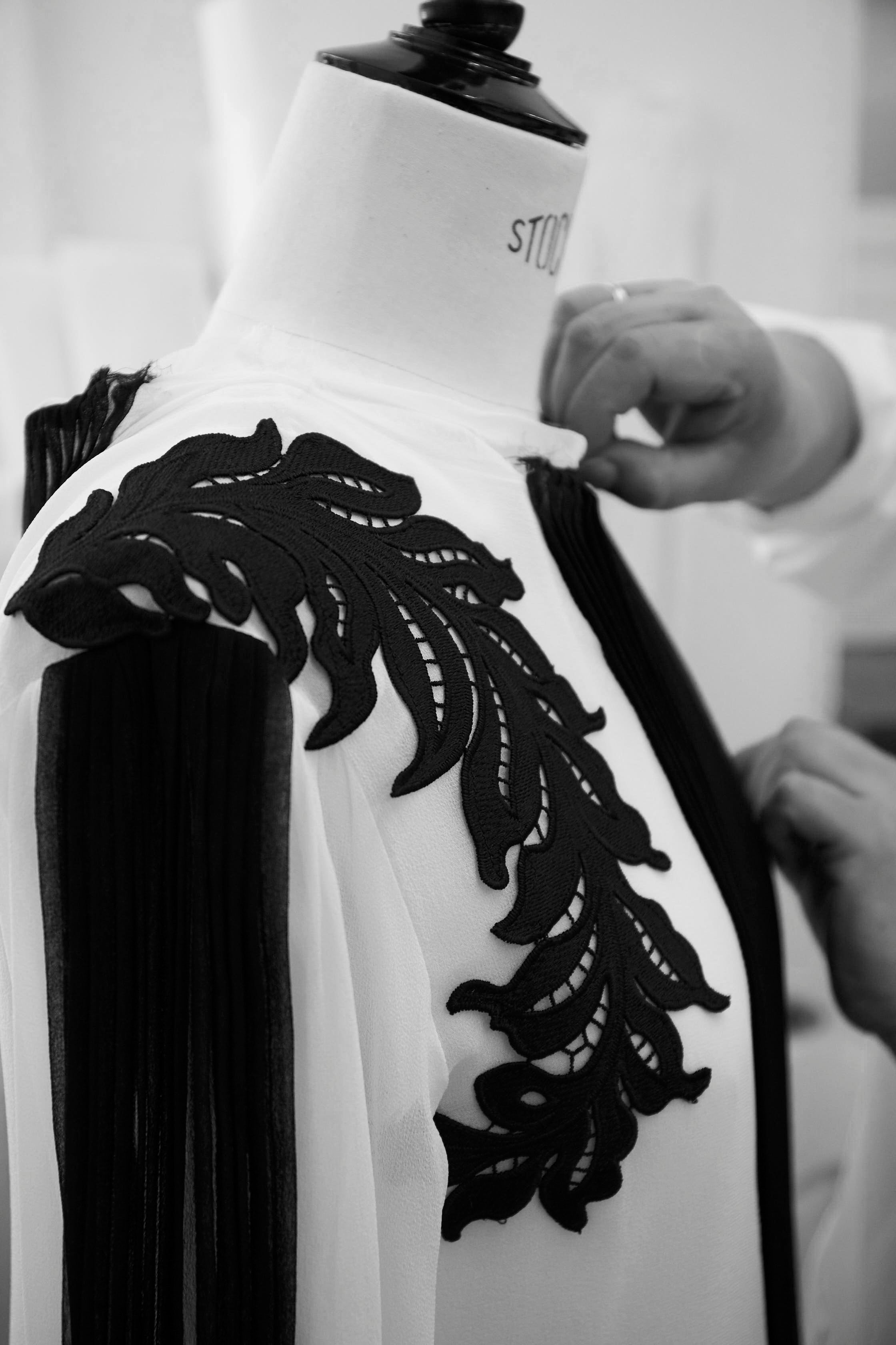 Close-up of a black lace applique being hand-stitched onto a white garment on a mannequin at the Costarellos Greek atelier