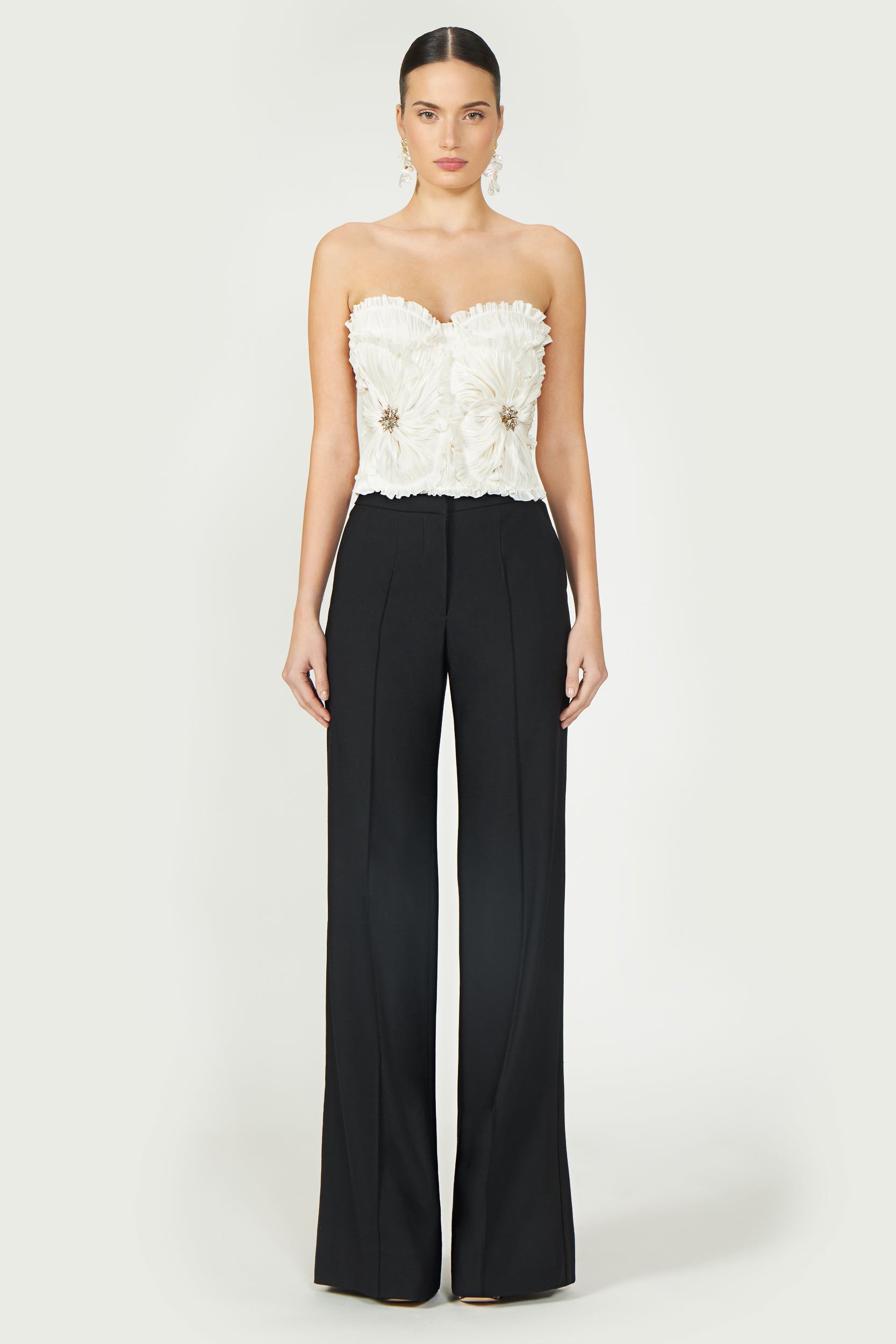 Model wearing Ezra Lurex Georgette Bustier paired with black trousers. Elegant off-white strapless top with floral pleats and jeweled accents.