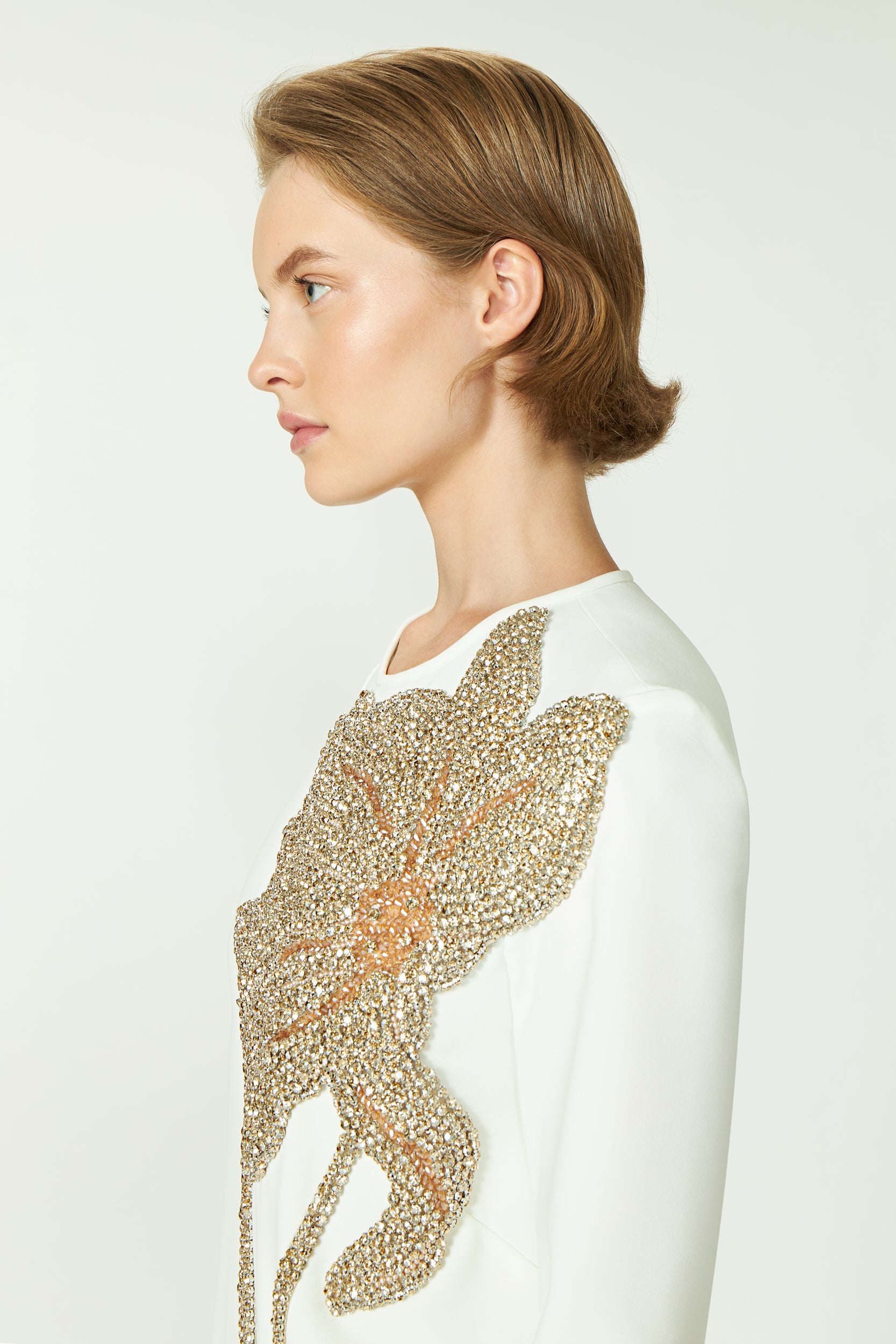 White top with gold sequin floral applique, side view, featuring round neckline and long sleeves, elegant and modern design.